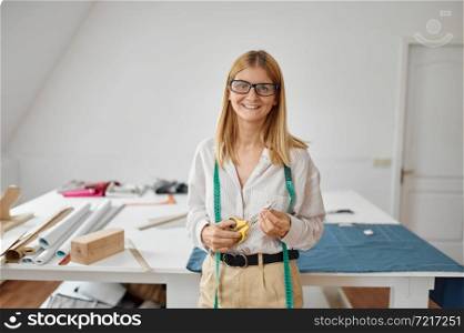 Smiling seamstress in glasses poses at her workplace in workshop. Dressmaking occupation and professional sewing, handmade tailoring business, handicraft hobby. Seamstress in glasses at her workplace in workshop