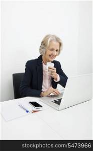 Smiling senior businesswoman having coffee while using laptop at desk in office