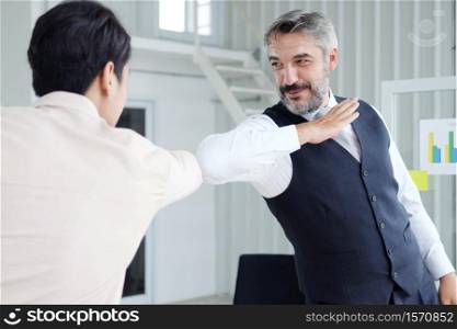 Smiling Senior Caucasian and young Asain businessman greeting with colleagues bumping elbows for preventing covid 19 virus in workspace at office. Social distance and new normal lifestyle concept.