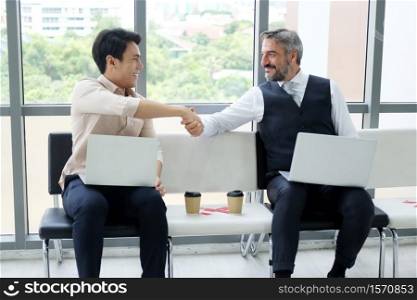 Smiling Senior Caucasian businessman shaking hand with young Asain businessman and working with laptop and sitting distance together in workspace at office. New normal lifestyle concept.