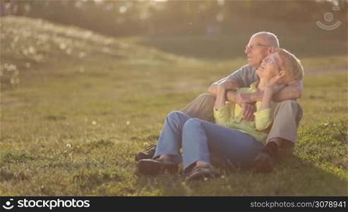 Smiling senior couple sitting on green grass in park and remembering funny momemnts of theis happy family life in glow of amazing sunset. Handsome elderly man embracing charming lady from behind while senior couple enjoying togetherness outdoor.