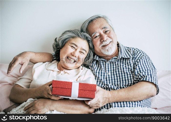 Smiling senior husband making surprise giving gift box to his wife in bedroom