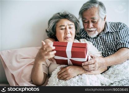 Smiling senior husband making surprise giving gift box to his wife in bedroom