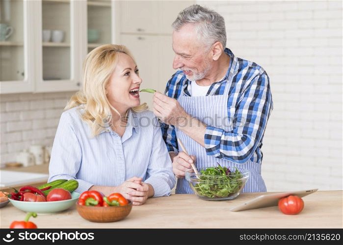 smiling senior man feeding fresh green salad her wife kitchen