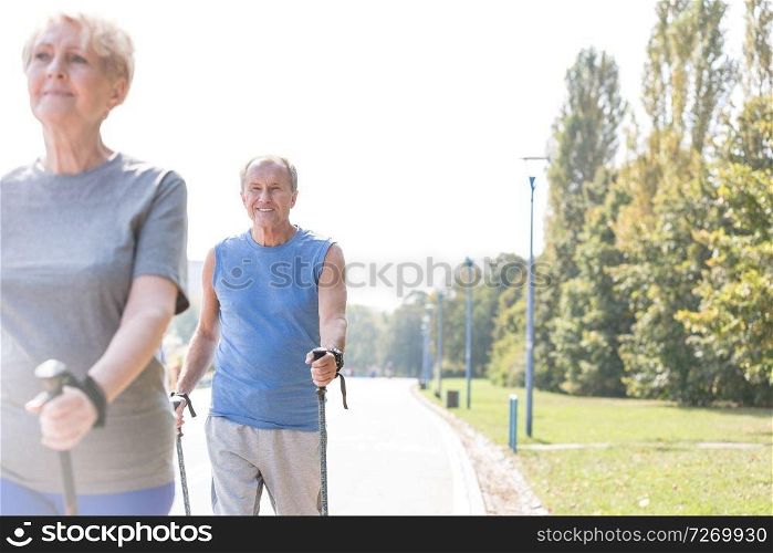 Smiling senior man using trekking poles while walking with woman in park