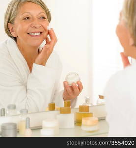 Smiling senior woman apply anti-wrinkles cream looking in bathroom mirror