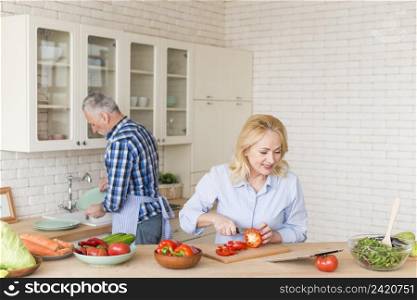smiling senior woman cutting red bell pepper with knife looking digital tablet his husband washing dishes kitchen sink