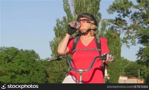 Smiling senior woman cyclist on bicycle drinking water from a plastic bottle, looking at camera