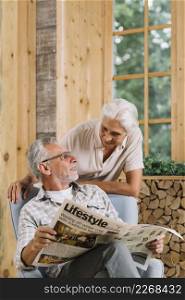 smiling senior woman looking her husband sitting chair holding newspaper