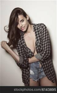 smiling sexy fashion brunette girl in pose with unbuttoned plaid shirt on nude breast and denim shorts. Erotic hipster style