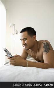 Smiling shirtless Asian young man lying on bed dialing or text messaging on cellphone.