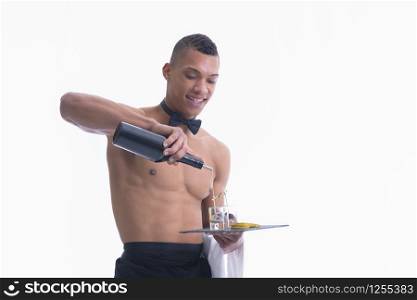 Smiling shirtless young waiter filling glass shots in a plate with liquor on a light background. Service and adult concept.. Shirtless young waiter filling glass shots with liquor