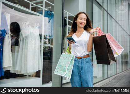 Smiling shopper young girl lifestyle standing using mobile phone after shopping on city street, Happy Asian beautiful woman holding shopping bag and smartphone on her hands near the mall shop window