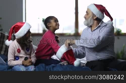 Smiling small mixed race girls reaching into santa hat held by joyful grandfather and picking up pieces of paper with player names during happy family playing secret santa on xmas eve. Joyful family spending leisure on winter holidays.