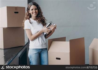 Smiling spanish female preparing moving to new home packing things in cardboard boxes. Glad woman holding stack of notebooks looking at camera on relocation day. Real estate sale, dwelling rental.. Smiling spanish female preparing moving to new home packing things in carton boxes on relocation day