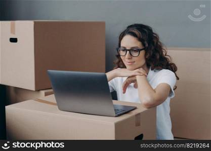 Smiling spanish girl in glasses looking at laptop screen, planning new home interior design, choosing furniture, shopping online, sitting with carton boxes. Relocation, apartment renovation concept.. Girl planning new home interior design at laptop near carton boxes. Relocation, apartment renovation