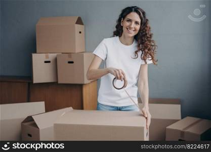 Smiling spanish woman packing stuff in cardboard box, using adhesive tape looking at camera. Female sealing carton package parcel with sticky tape, preparing for relocation. Delivery, removal service.. Smiling spanish woman packing stuff in cardboard box, using adhesive tape. Delivery, removal service