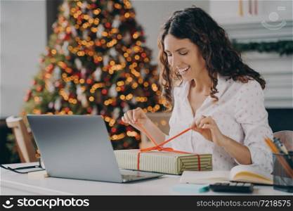 Smiling spanish woman unpacking Christmas gift while sitting at work desk with laptop, happy female opening wrapped box and feeling excited during remote job at home decorated for xmas holidays. Smiling woman unpacking Christmas gift while sitting at work desk with laptop against xmas tree