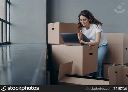 Smiling spanish woman with cardboard boxes works at laptop searching moving company online, shopping furniture for new apartment, planning home renovation. Ecommerce, relocation day concept. Female with boxes works at laptop, orders moving company, shopping online. Ecommerce, relocation day