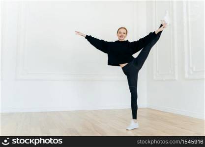 Smiling sporty female stands on one leg in fitness center, demonstrates nice physical resilience, has slim body shape, dressed in black hoody and leggings, wears white sneakers. People and balance