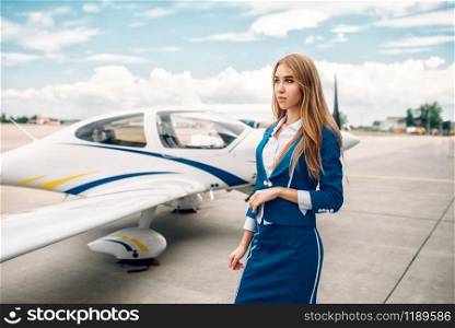 Smiling stewardess in uniform poses against small airplane. Air hostess in suit near plane. Private airline, flight attendant. Smiling stewardess in suit against small airplane