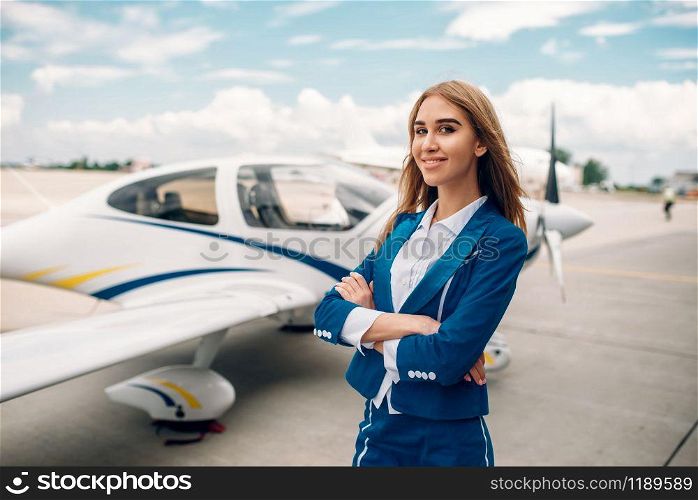 Smiling stewardess in uniform poses against small airplane. Air hostess in suit near plane. Private airline, flight attendant. Smiling stewardess in suit against small airplane