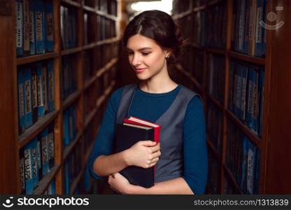 Smiling student girl or woman with books between bookshelves in library.. Student girl with books between bookshelves