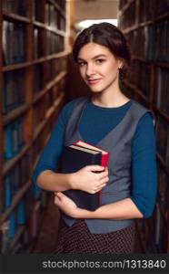 Smiling student girl or woman with books between bookshelves in library.
