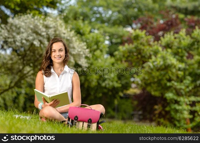 Smiling student girl sitting on grass with open book summer