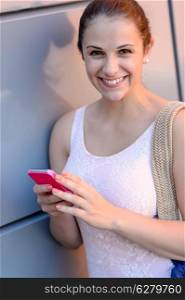 Smiling student girl with mobile phone leaning against modern wall