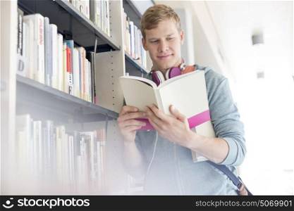 Smiling student reading book by shelf at library in university