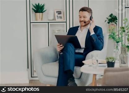 Smiling stylish businessman in formal wear sitting in armchair and looking at laptop screen while chatting online and using headset, sales manager having online meeting during remote job at home. Happy young businessman in suit having pleasant online conversation on laptop