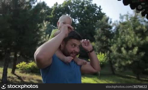 Smiling stylish father with beard carrying his adorable toddler son on shoulders while walking in asian styled park on sunny summer day. Cheerful infant child with blue eyes riding his dad&acute;s shoulders and smiling as they spend leisure together.