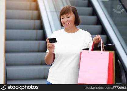 Smiling stylish middle aged woman with credit, debit card and shopping bags standing on shopping mall background. Autumn holidays, discounts, black Friday. concept of consumerism, sale, rich life.. Smiling stylish middle aged woman with credit, debit card and shopping bags standing on shopping mall background. Autumn holidays, discounts, black Friday. concept of consumerism, sale, rich life