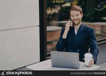 Smiling successful business owner working on laptop remotely and talking with employee on mobile phone, wearing formal suit, sitting in sidewalk cafe and making phone call, holding white coffee cup. Young successful business owner working on laptop in cafe outside and talking on mobile phone