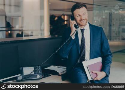 Smiling successful businessman in suit talking on phone sitting in office on top of desk, holding laptop and agenda, answering office call, checking implementation of recommendations and daily reports. Smiling businessman talking on phone sitting in office on top of desk, holding laptop and agenda