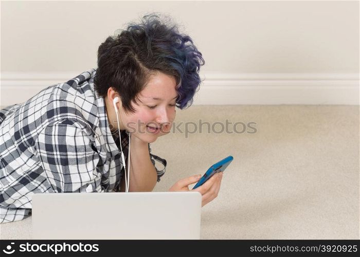 Smiling teen girl looking at cell phone with computer in forefront while lying down listening to music at home.