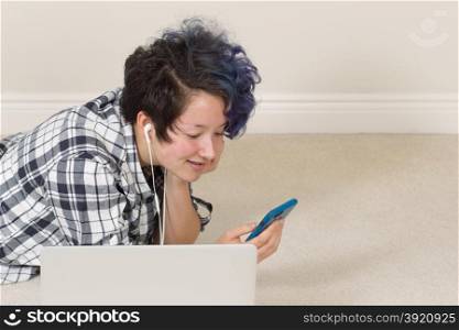 Smiling teen girl looking at cell phone with computer in forefront while lying down listening to music at home.