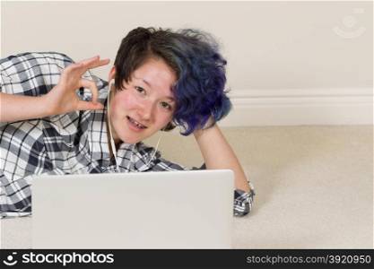Smiling teen girl, looking forward, giving okay sign while using computer and listening to music at home.
