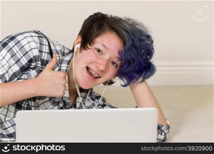 Smiling teen girl, looking forward, giving thumbs up while using computer and listening to music at home.