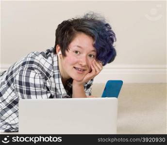 Smiling teen girl, looking forward, with cell phone in hand with computer in forefront while lying down listening to music at home.