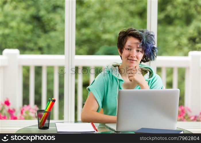 Smiling teen girl, looking forward, with computer, books and pencils in forefront. Large windows in background with blurred out bright green trees and flowers.