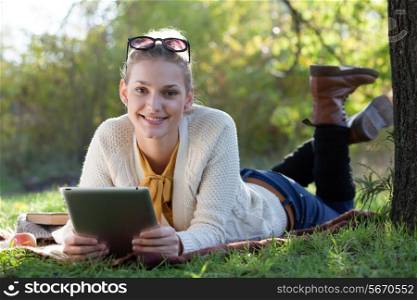 smiling teen girl lying with tablet pc under the tree in evening sunlight outdoors
