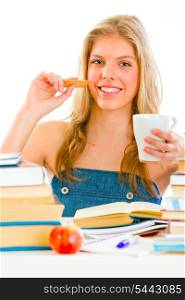 Smiling teen girl sitting at table with books and drinking tea with cookie&#xA;