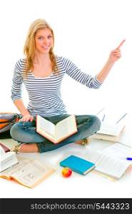 Smiling teen girl sitting on floor among schoolbooks and pointing in corner isolated on white &#xA;