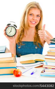 Smiling teen girl with alarm clock sitting at table and showing thumbs up gesture&#xA;