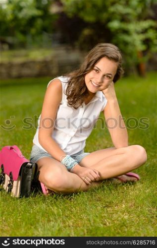Smiling teenage girl sitting on grass with pink satchel summer