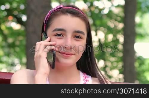 Smiling teenager girl talking on smartphone in city park.