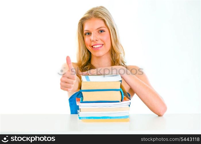 Smiling teengirl sitting at table with books and showing thumbs up gesture&#xA;