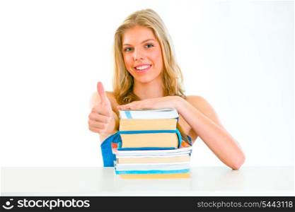 Smiling teengirl sitting at table with books and showing thumbs up gesture&#xA;